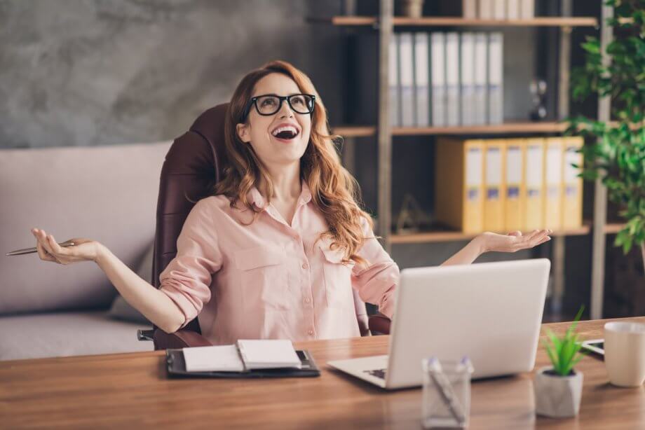 Photograph of a woman with glasses & wavy red hair sitting in an office chair seemingly happily surprised